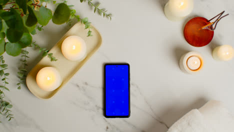 Overhead-View-Looking-Down-On-Still-Life-Of-Blue-Screen-Mobile-Phone-Lit-Candles-And-Incense-Stick-With-Green-Plant-And-Towels-As-Part-Of-Spa-Day-Decor-2